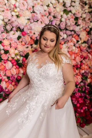 Plus-Size-Braut in einem Spitzenkleid vor einer Blütenwand